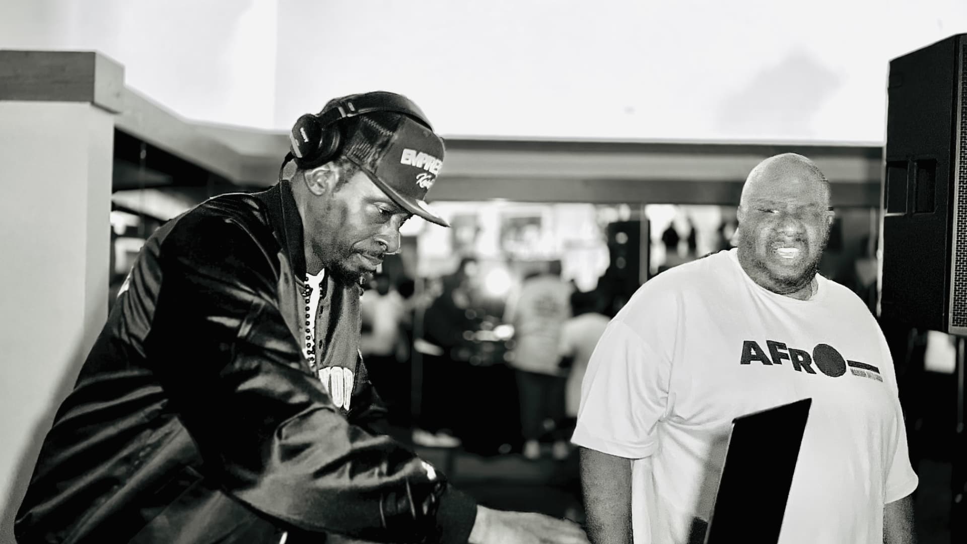 A DJ wearing headphones at the decks with another man smiling beside him