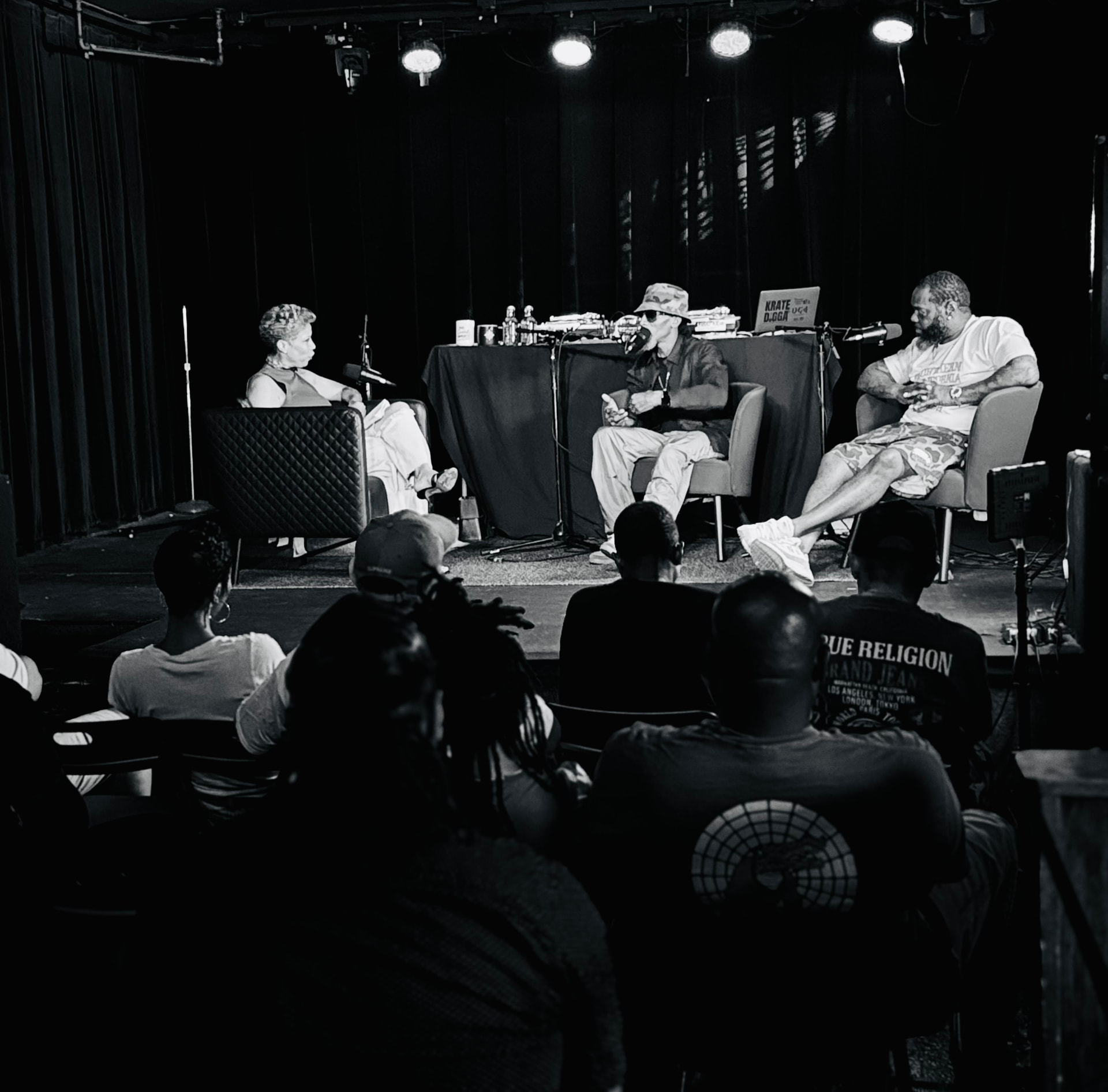 Black-and-white photo of three panelists seated in armchairs on stage during a live recording, seen from the audience