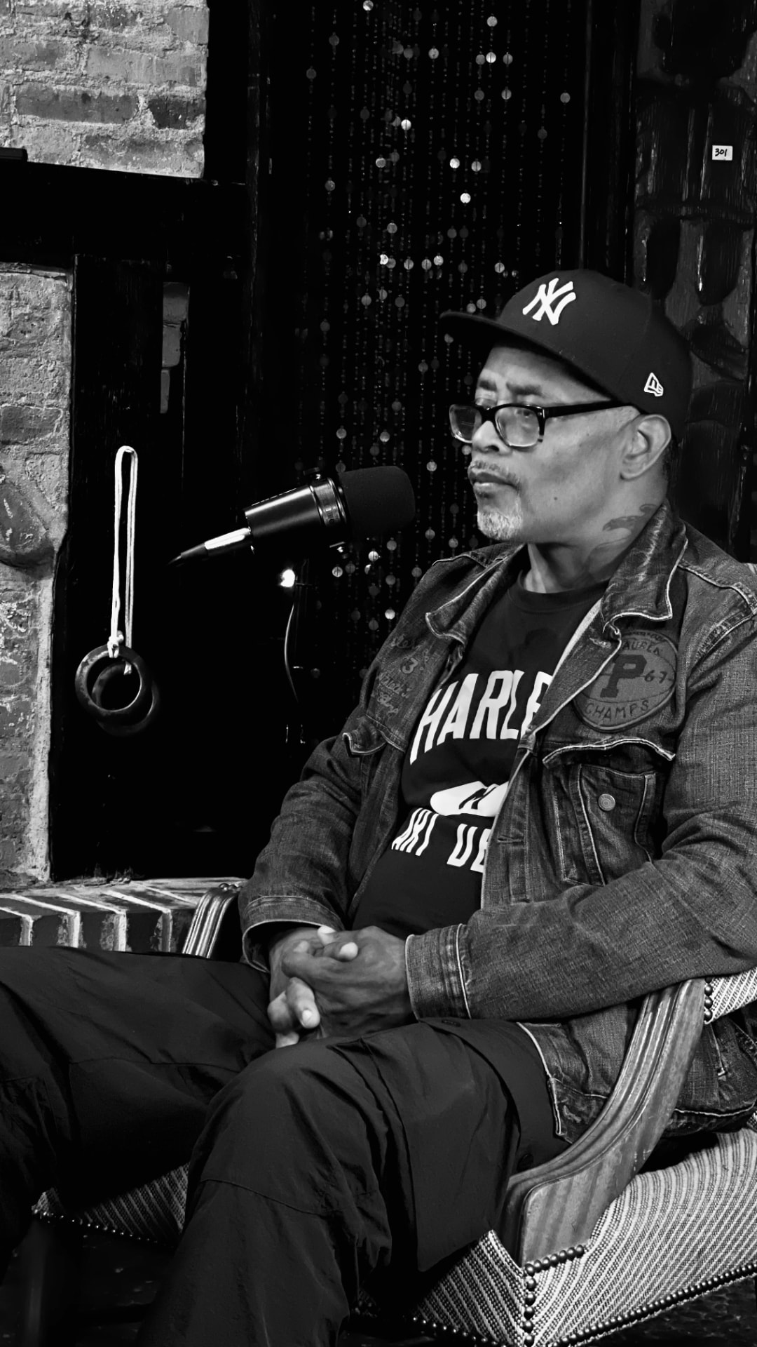 A man in a cap and denim jacket seated in a studded armchair in front of a boom microphone during a studio interview
