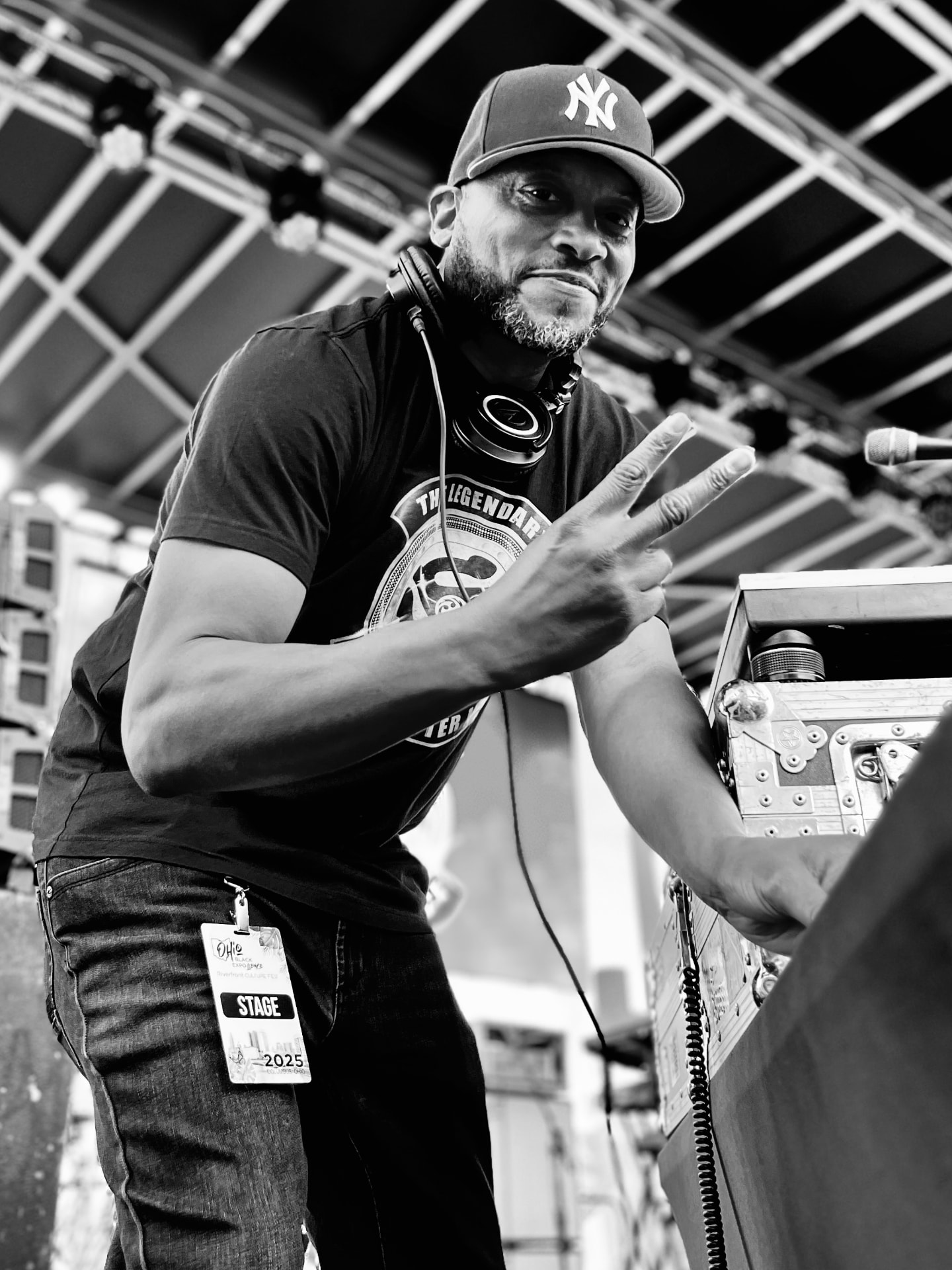 A DJ in a New York cap with headphones around his neck making a peace sign beside his equipment