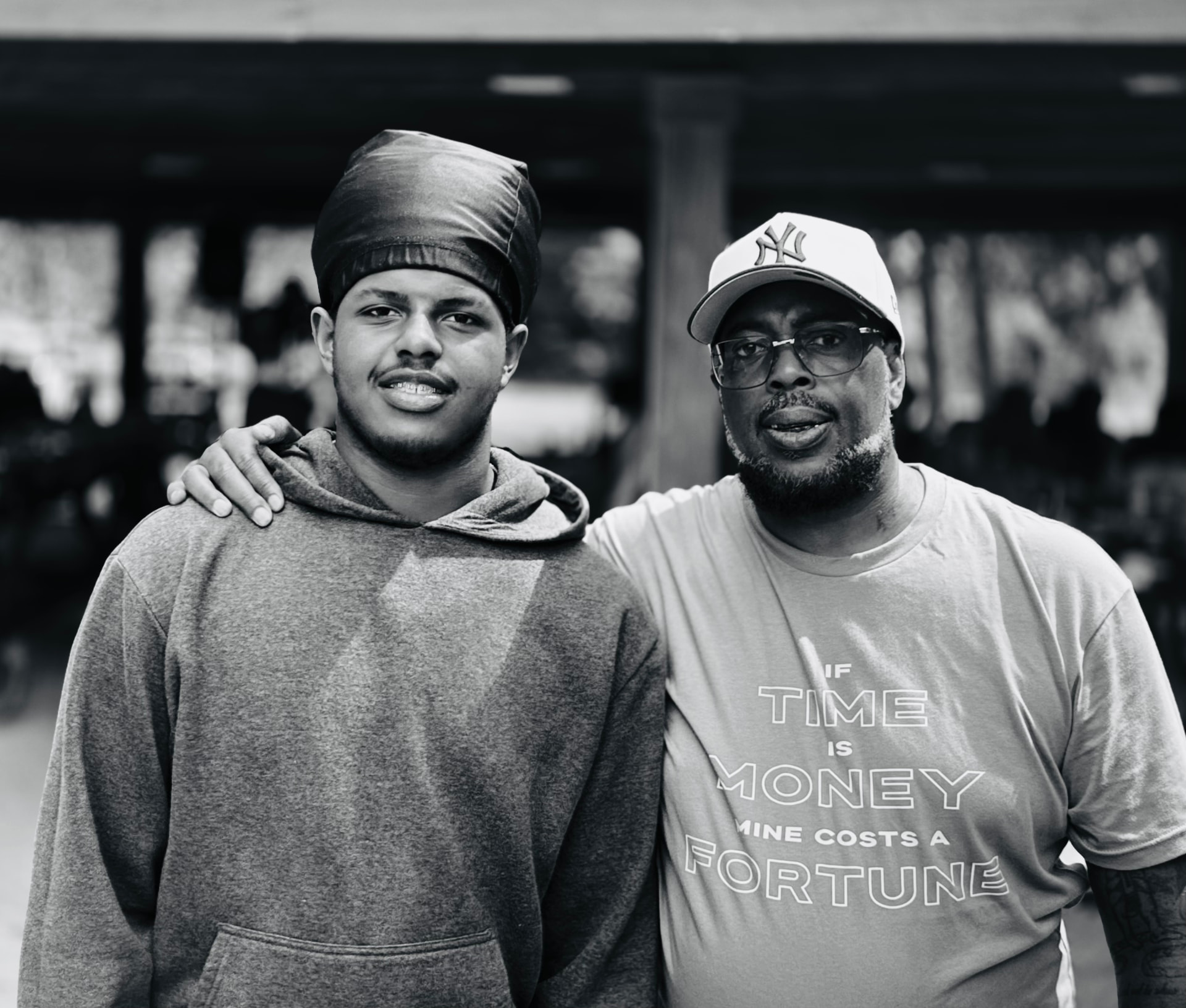 Two men standing arm in arm under an open-air pavilion, one in a hooded sweatshirt and headwrap, the other in a light cap and a graphic T-shirt reading “If time is money, mine costs a fortune”