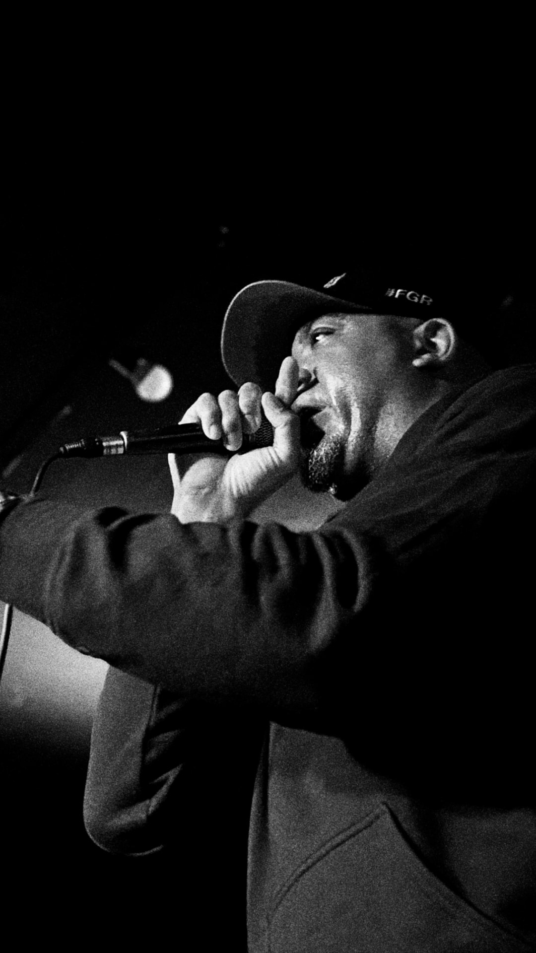 A performer in a dark hoodie and a cap reading #FGR gripping a wired microphone on a dark stage, lit by a single round stage light