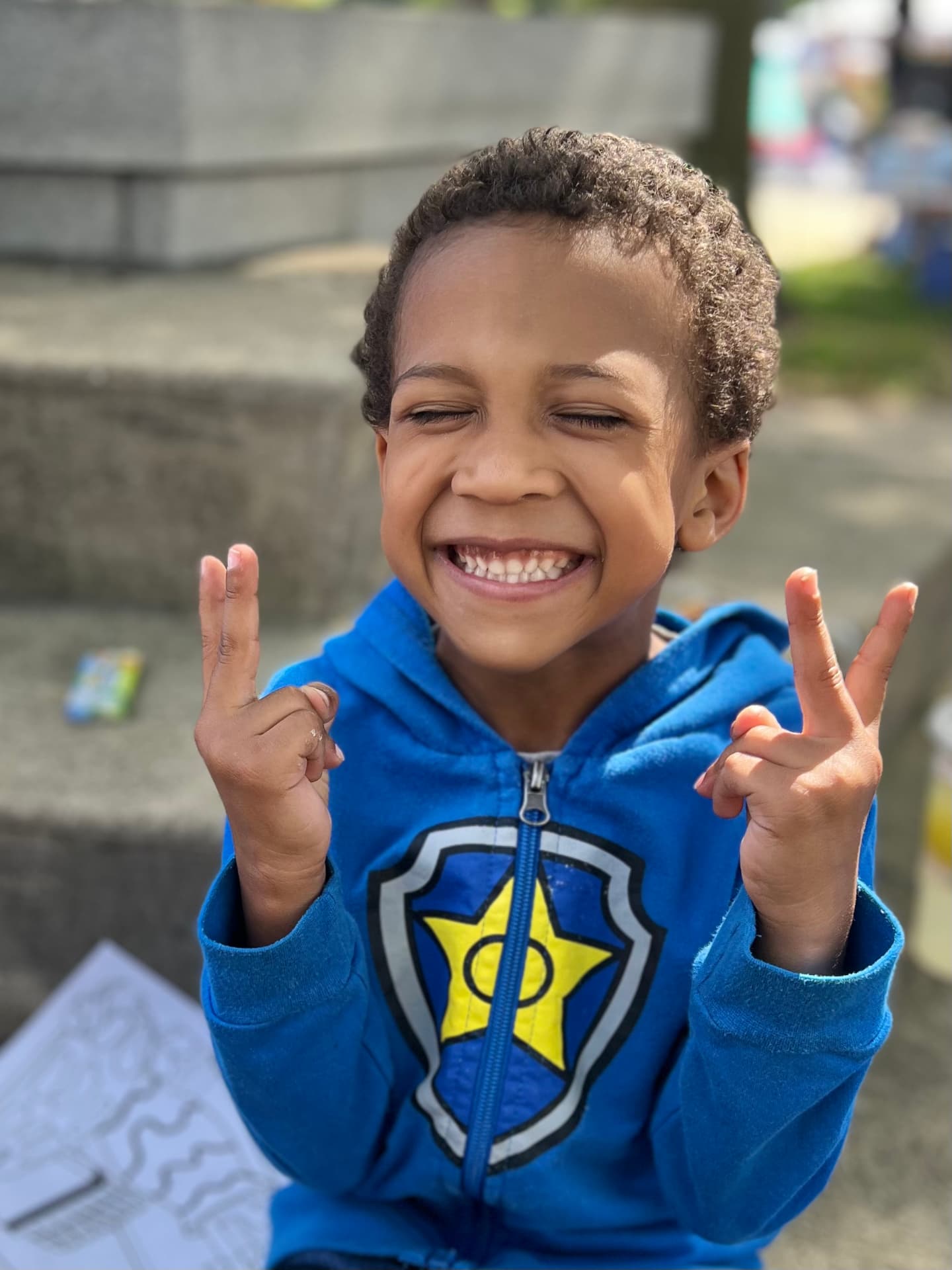 A smiling boy in a blue hooded shirt making peace signs with both hands