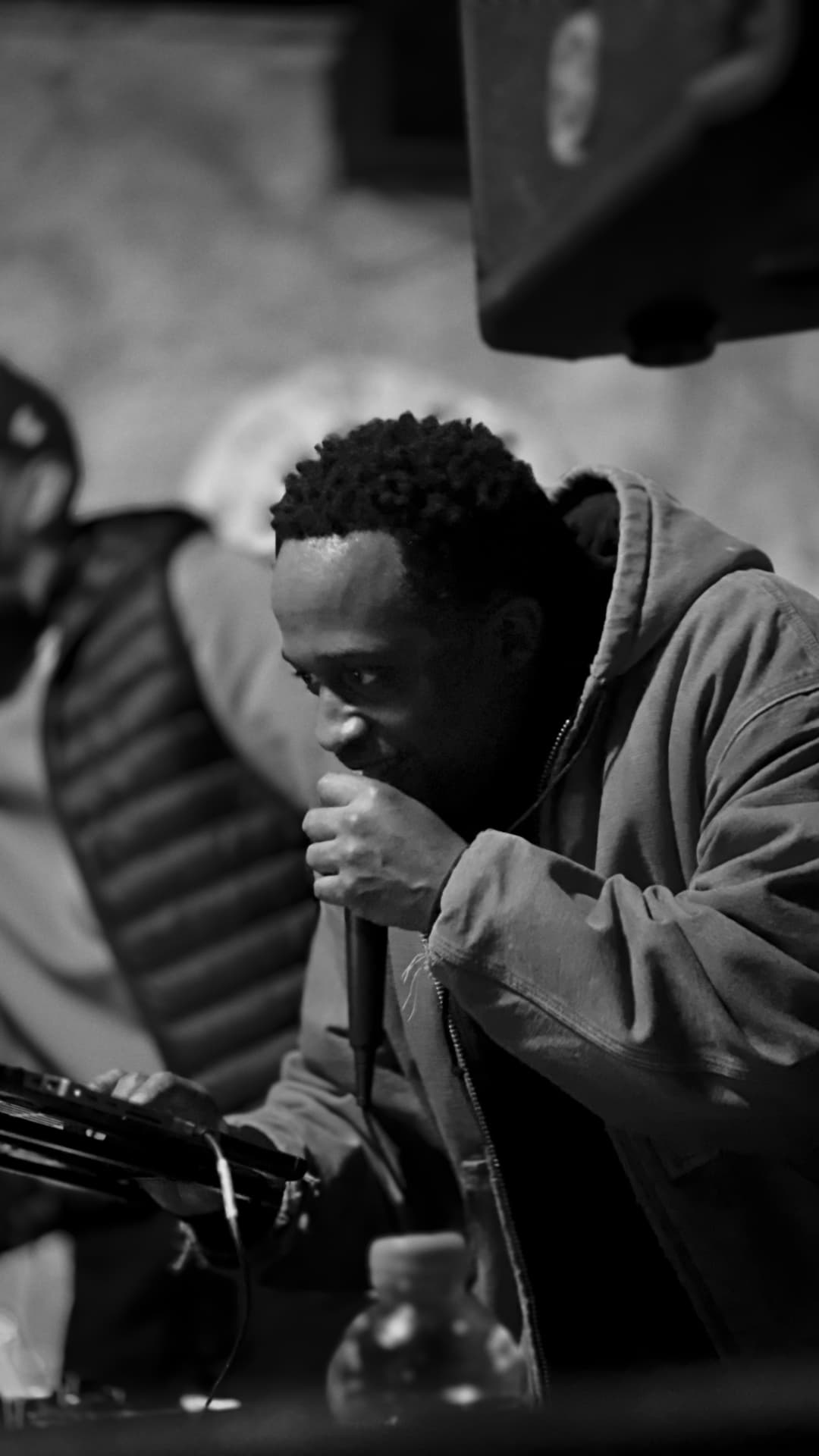 A man in a hooded jacket leaning into a microphone in low light