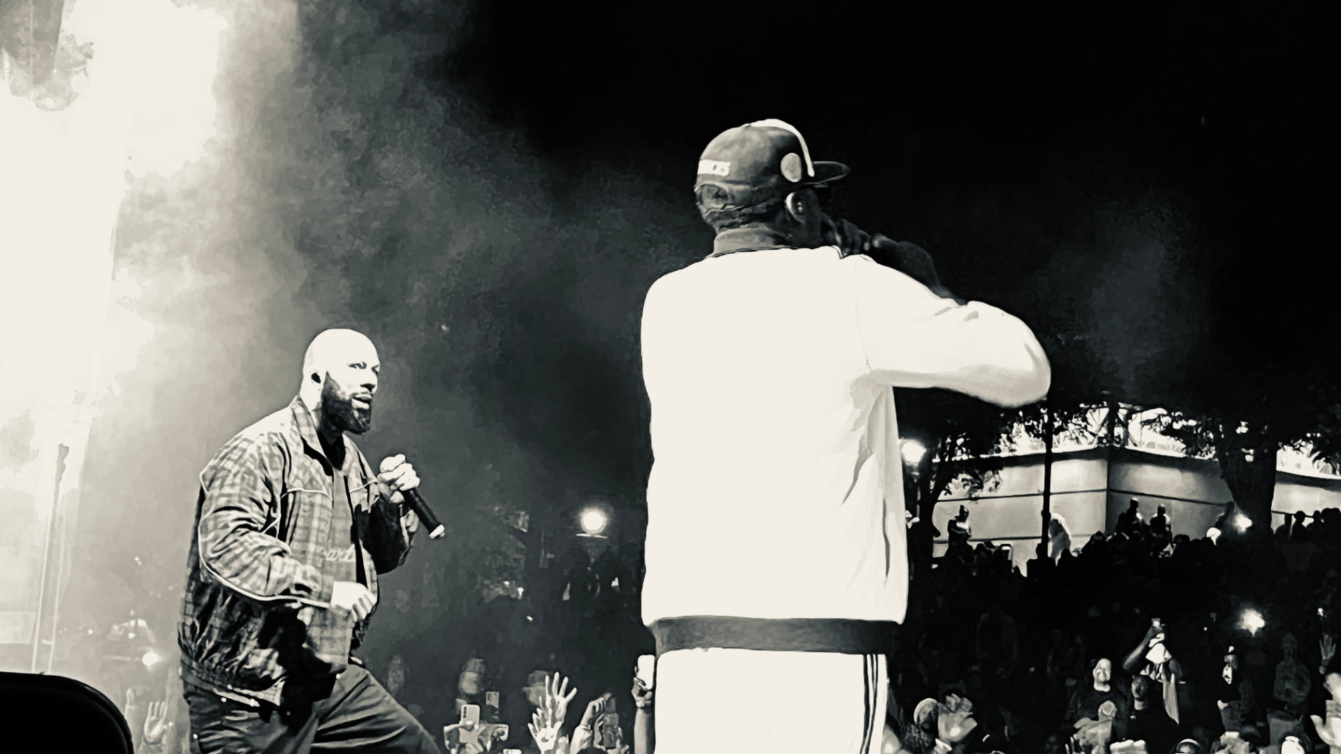 A bearded performer in a plaid jacket singing into a microphone on an outdoor night stage, seen past a crew member in a cap, with a crowd raising phones below