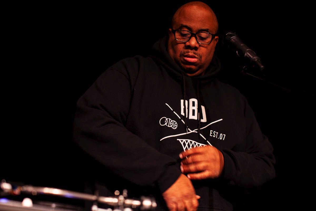 A bald man in dark-framed glasses and a black hoodie at a turntable, a microphone stand behind him on a dark stage