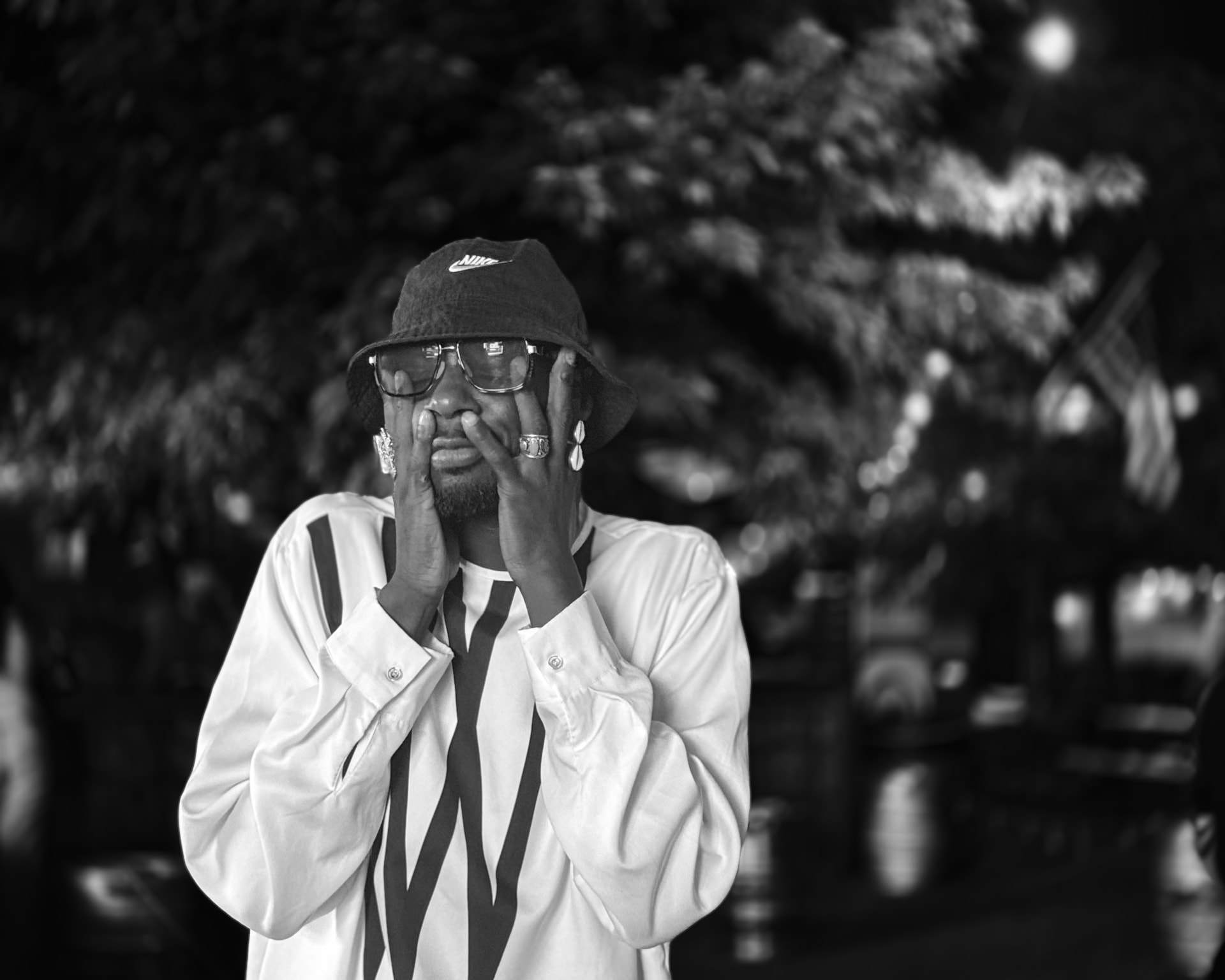 A man in a bucket hat and glasses framing his face with both hands, wearing rings, against blurred evening trees