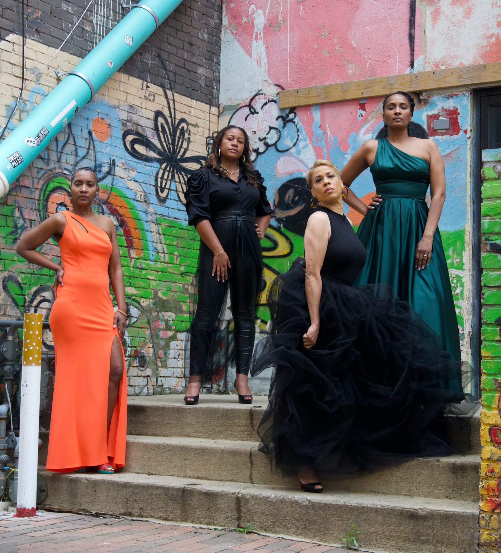 Four women in formal gowns posing on concrete steps in front of a painted mural wall