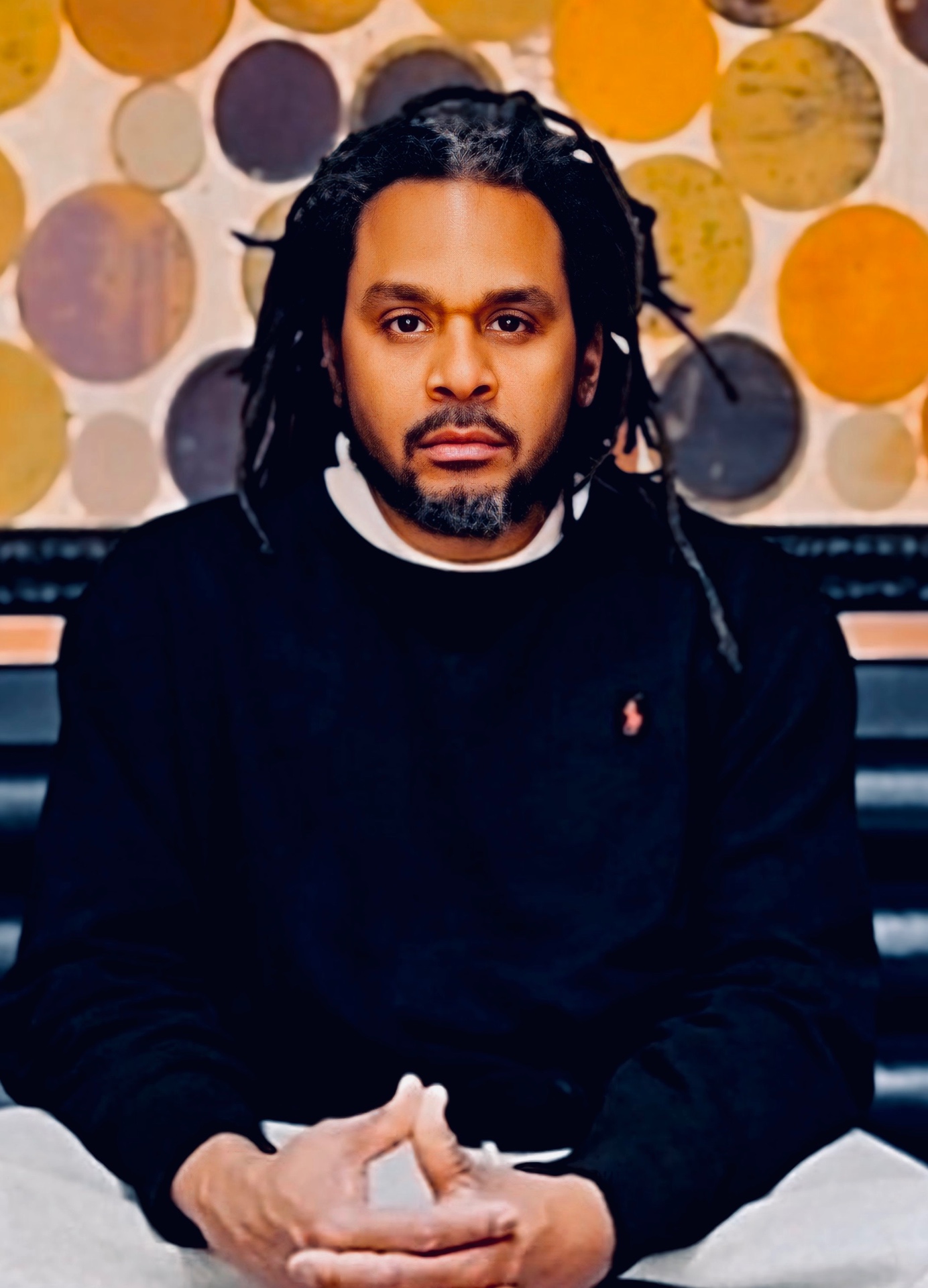 Charles Ewing seated on a black bench in front of an artwork of circular discs in gold, mauve and dark brown