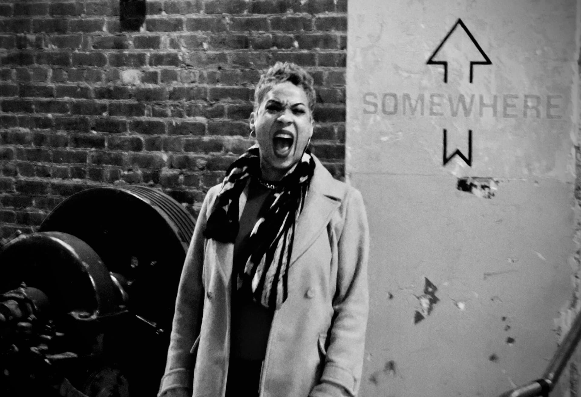A woman cheering in front of a brick wall and a sign reading SOMEWHERE