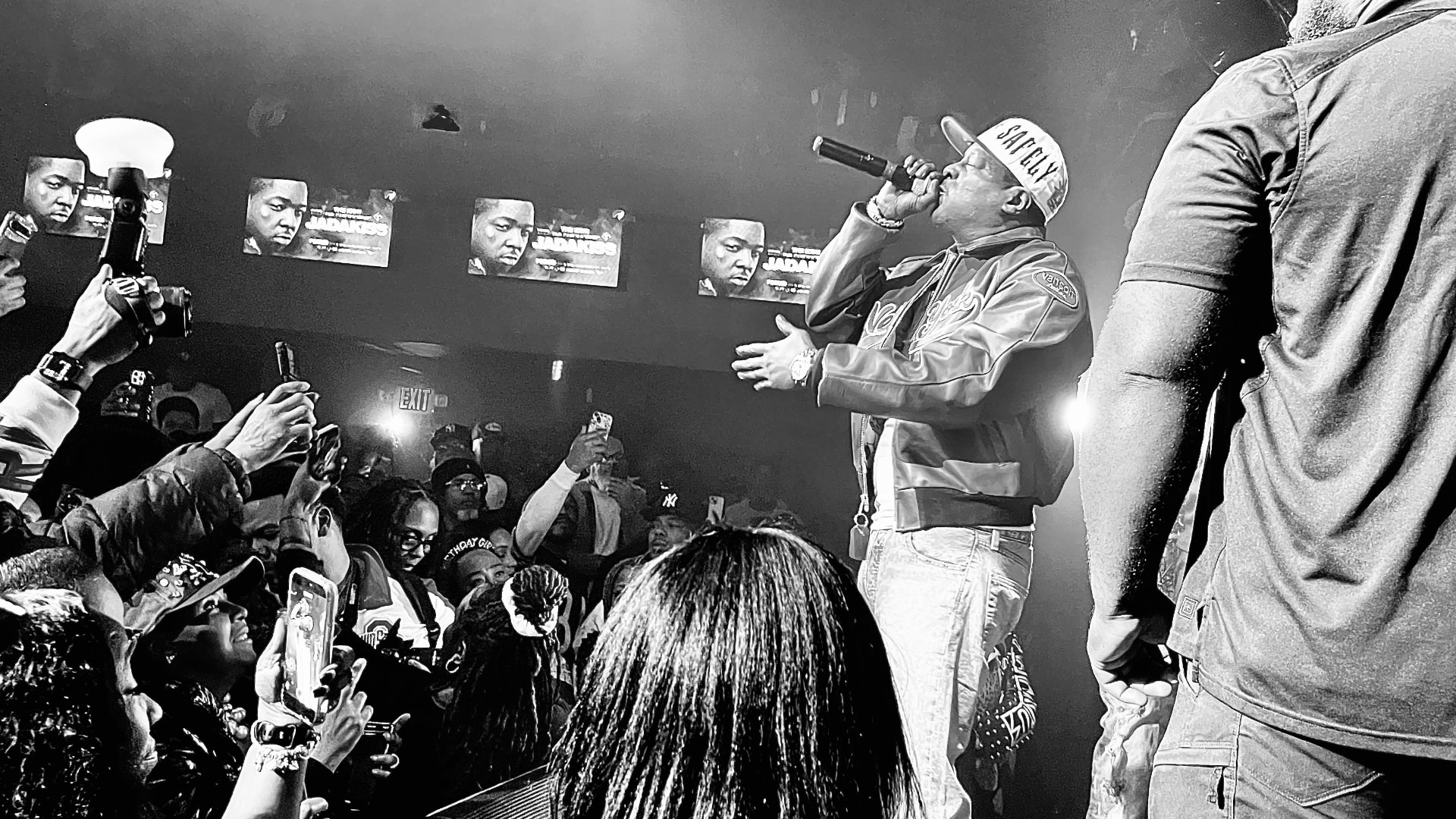 A performer in a cap and jacket singing into a handheld microphone on a stage, seen past a crowd raising phones below, with lit screens along the wall behind