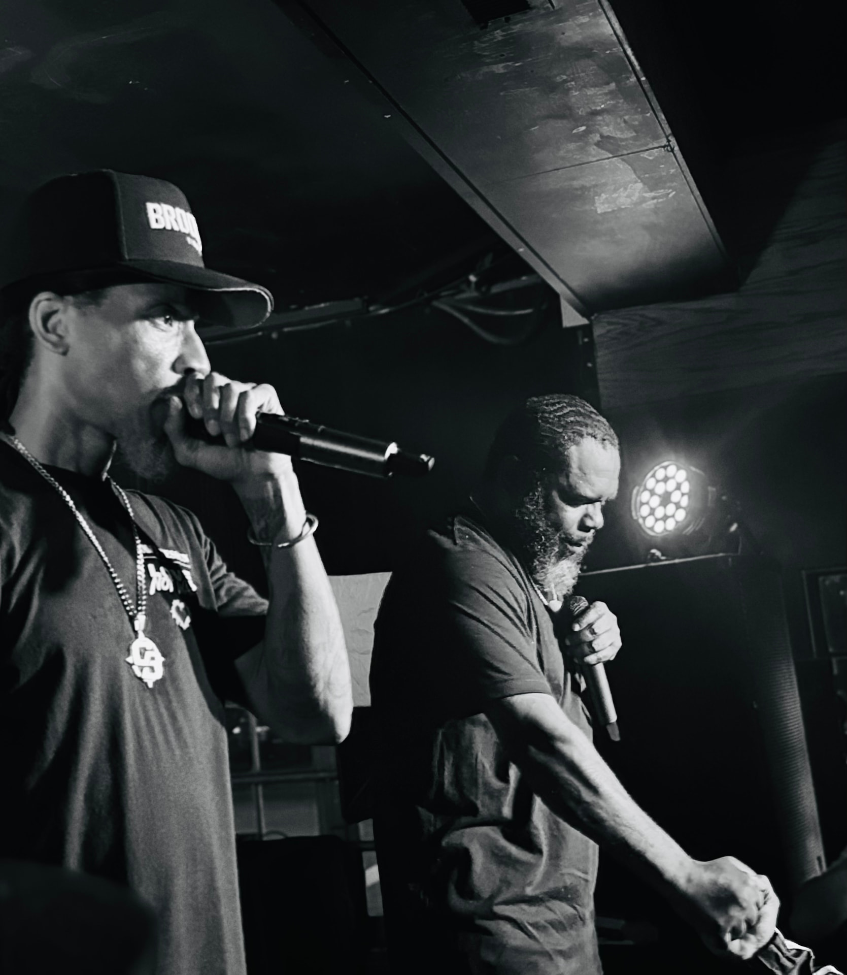 Two performers on a dimly lit stage, one wearing a Brooklyn cap, lit by a round stage light