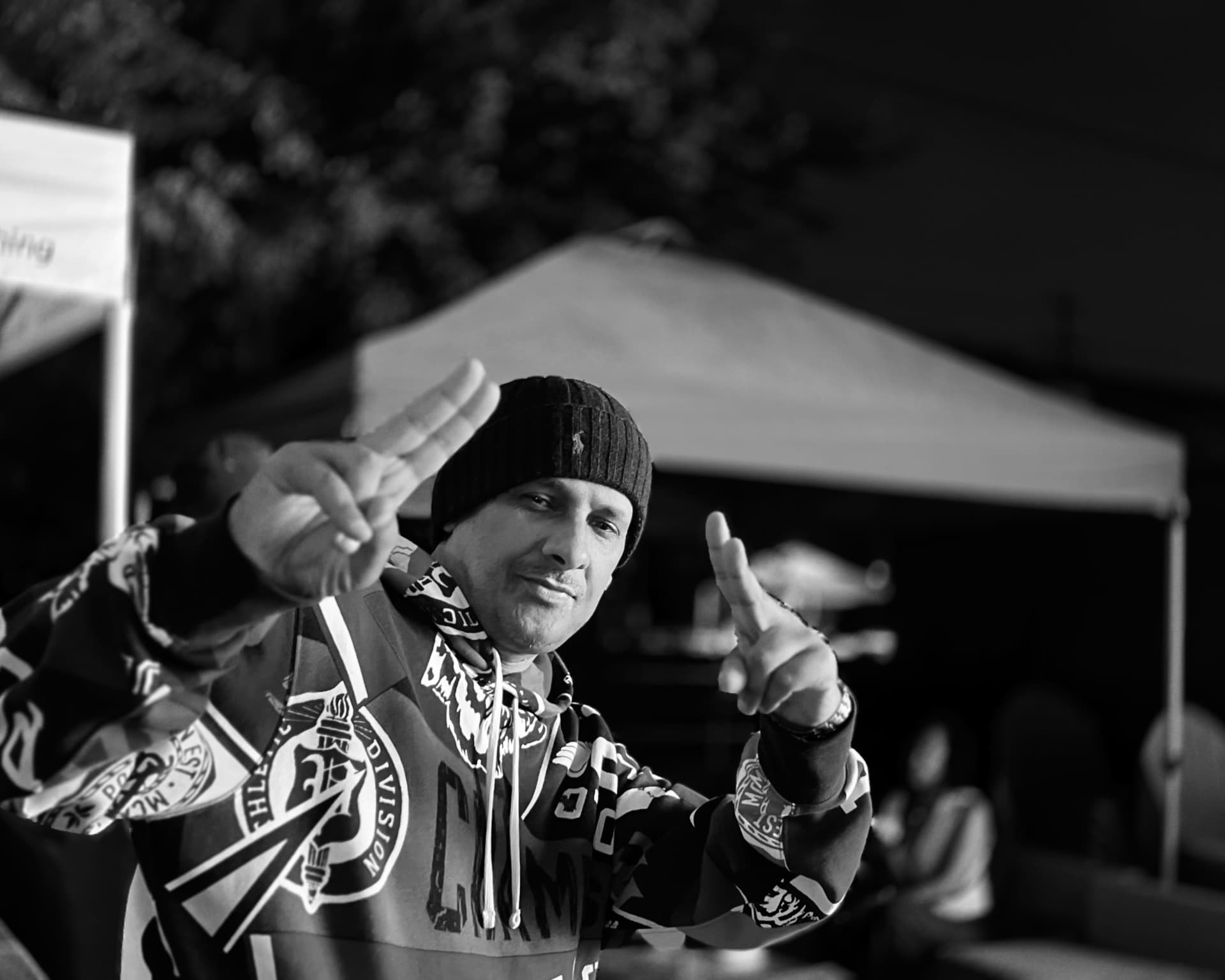 A man in a beanie and hoodie making hand gestures toward the camera at an outdoor night gathering