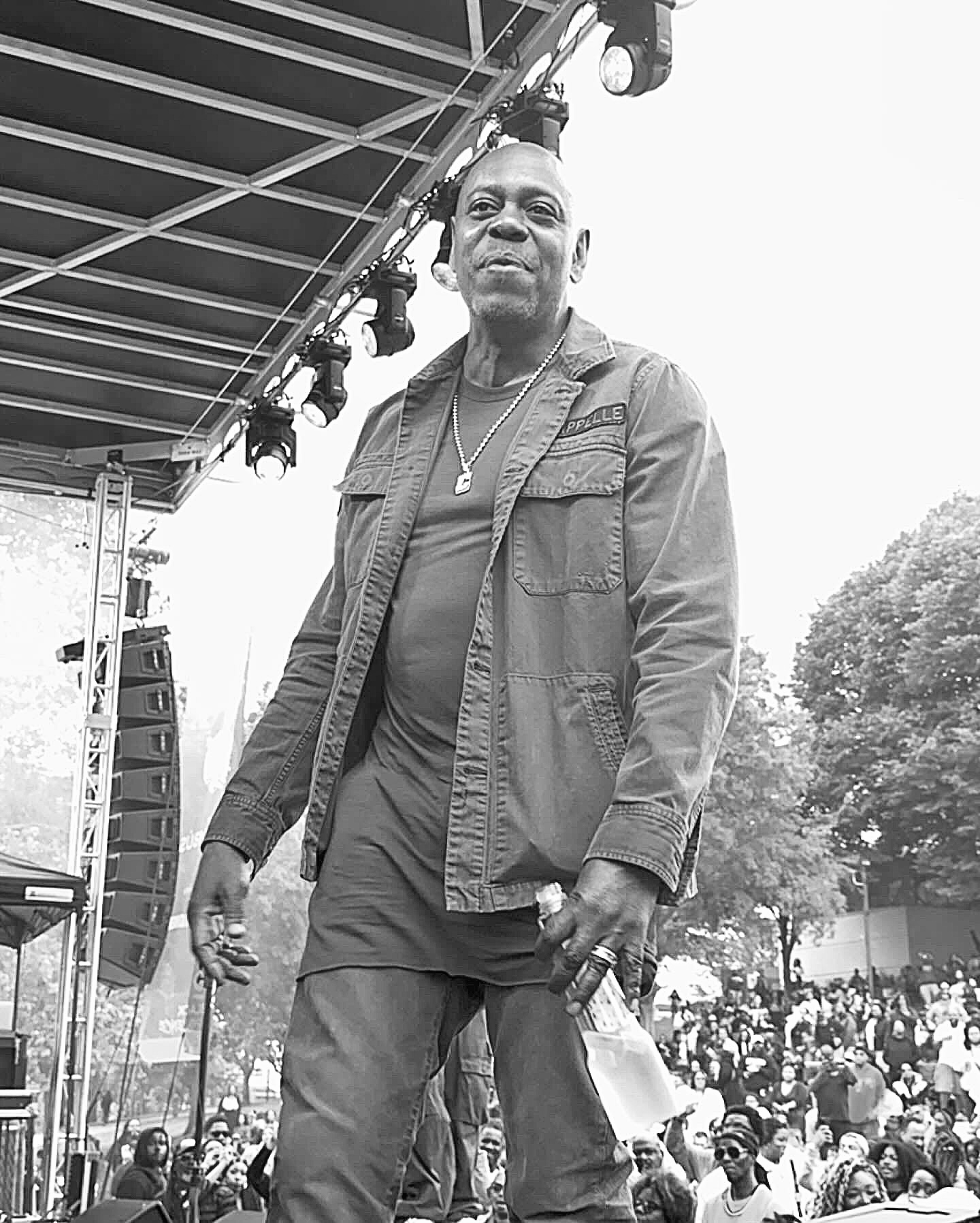A man in a denim jacket holding a water bottle on an outdoor festival stage, a crowd behind him