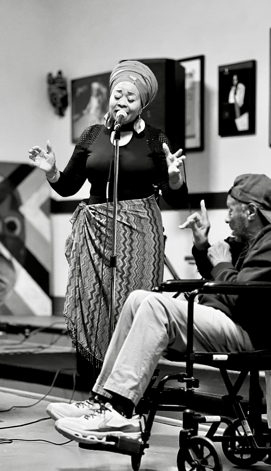 A woman in a headwrap singing into a stand microphone while a man seated in a wheelchair gestures beside her