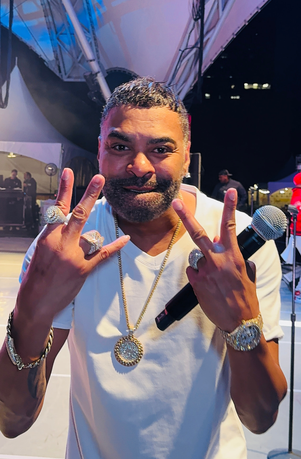 A close-up of a man in a white shirt with jewelry and a microphone making hand signs backstage