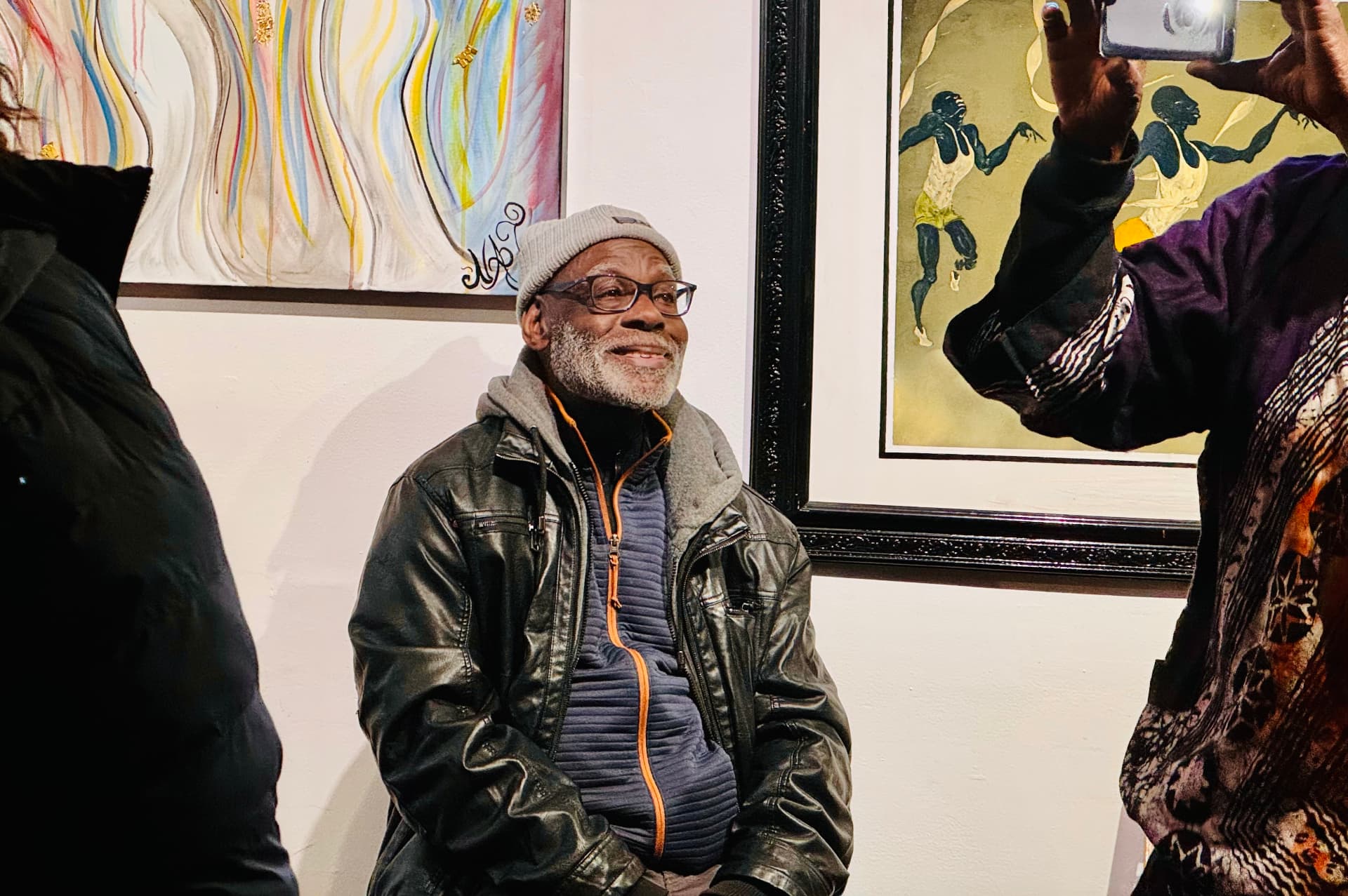 An older man in a beanie and glasses smiling beneath framed artwork at an art-gallery event
